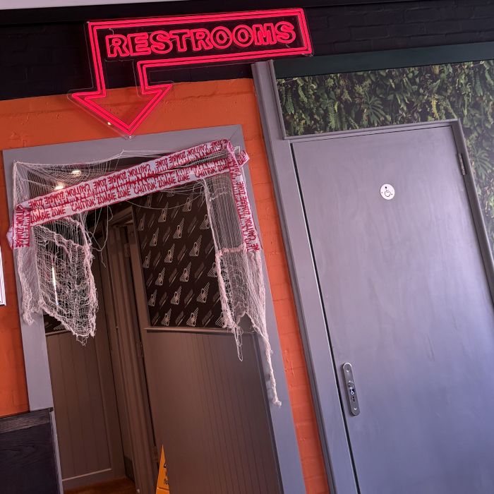 Custom red LED neon 'Restrooms' sign with a downward arrow above a doorway, decorated with cobwebs and zombie caution tape