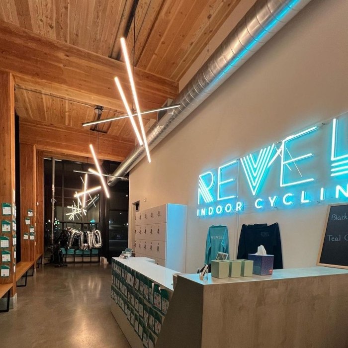 Reception area of Revel Indoor Cycling with a blue custom neon logo sign on the wall and linear pendant lighting