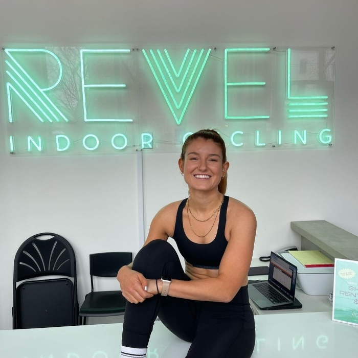 Woman in black activewear sitting on the reception desk in front of a green custom neon 'Revel Indoor Cycling' sign