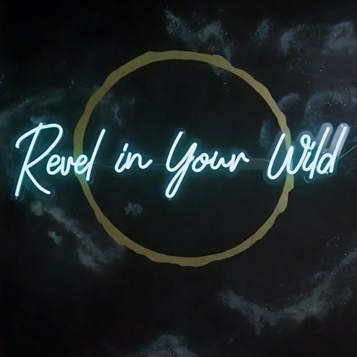 Revel in Your Wild ice blue script LED neon sign inside a gold circle on a dark celestial mural