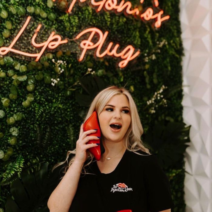 Team member with a red stiletto phone beneath the coral San Antonio's Lip Plug script LED neon sign on a greenery wall