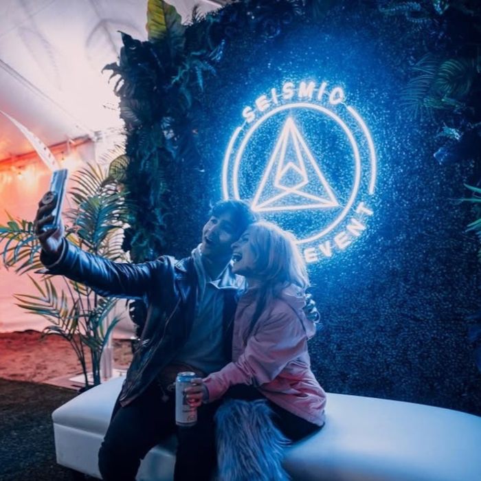 Couple taking a selfie in front of a blue Custom Neon® 'Seismic Event' triangle logo sign on a greenery event wall