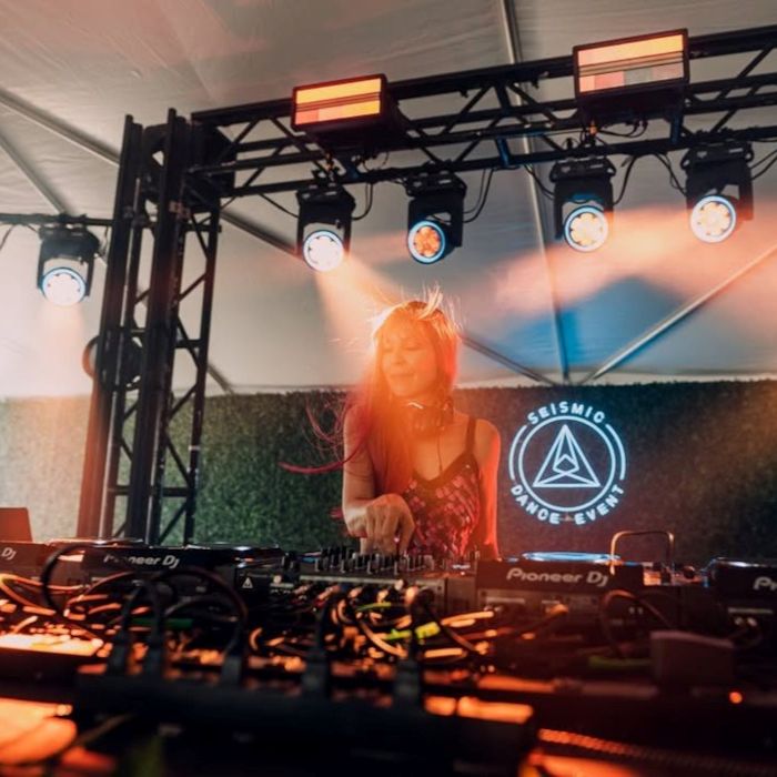 Female DJ performing under stage lights at a Seismic Dance Event booth in front of a white Custom Neon® triangle logo sign on a hedge wall