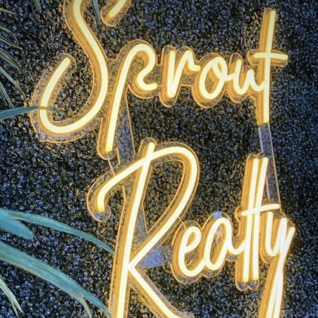 Warm gold custom neon 'Sprout Realty' script sign on a black foliage greenery wall with palm leaves