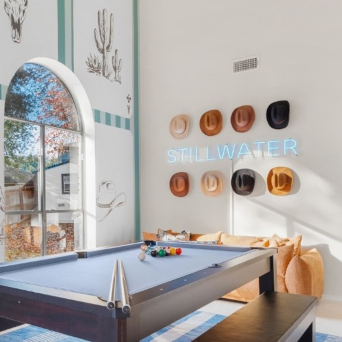Ice blue STILLWATER LED neon sign framed by a grid of cowboy hats above the pool table in a vacation home designed by Somerled