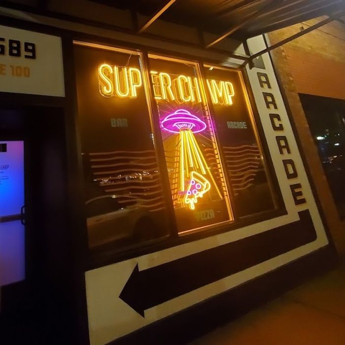 Nighttime storefront window with an orange custom neon 'Superchamp' bar and arcade sign and a pink neon UFO beaming up a pizza slice