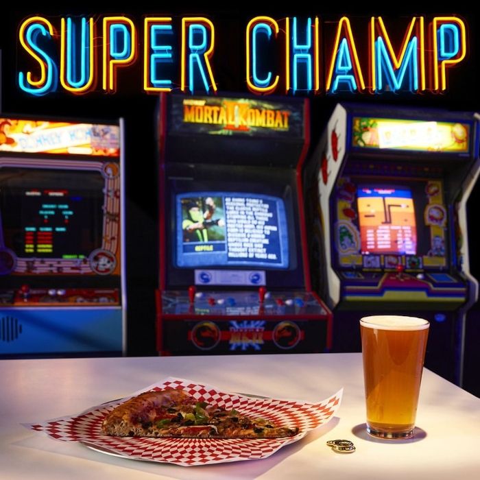 Blue and orange custom neon 'Super Champ' sign above vintage arcade machines, with a pizza slice and a pint of beer on a table