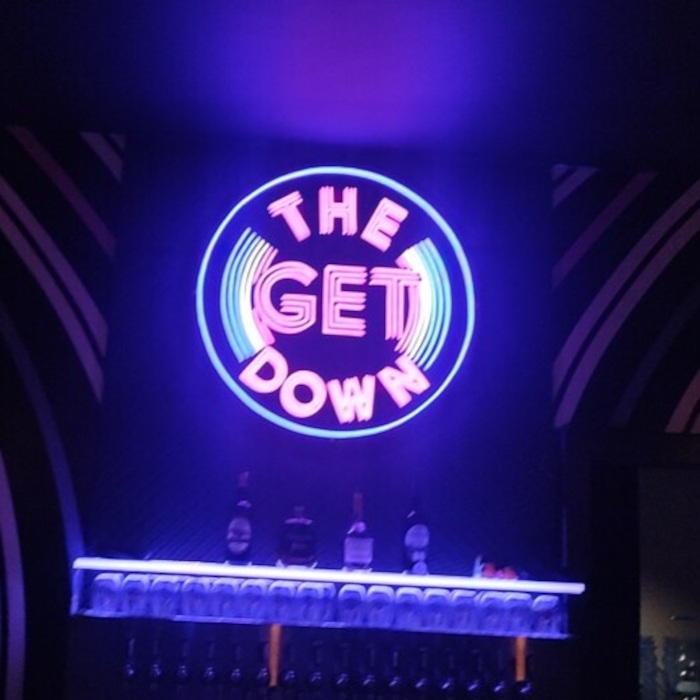 Purple, pink and blue custom neon 'The Get Down' circular record logo sign glowing on a dark wall above a bar