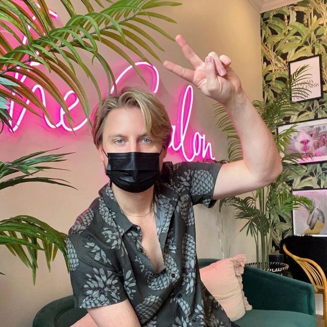 Drew Storin making a peace sign in front of a pink LED neon salon sign among palm plants