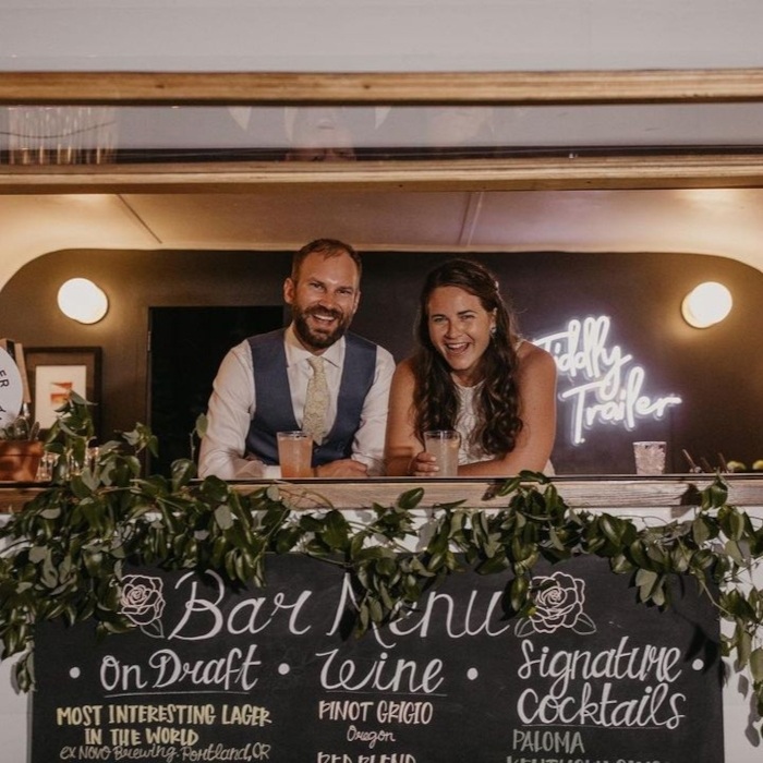 Bride and groom leaning out of a vintage caravan bar window with a pink custom neon 'Tiddly Trailer' sign, above a chalkboard drinks menu