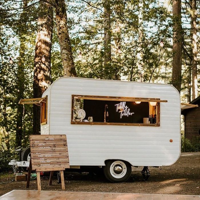 Vintage white caravan mobile bar in the woods with a pink custom neon 'Tiddly Trailer' sign glowing in the window