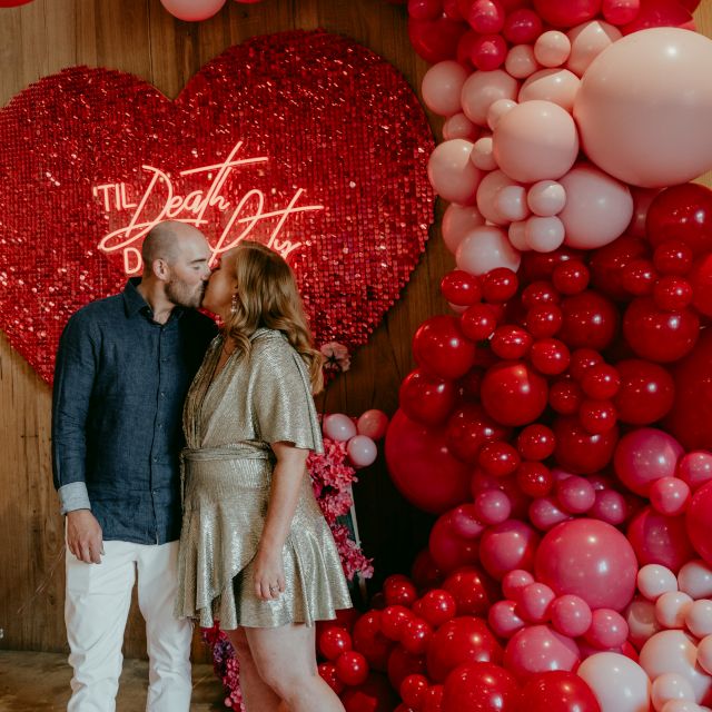 Red Til Death Do Us Party Custom Neon® sign on a red shimmer backdrop surrounded by florals and a red balloon waterfall @thewarehousegeelong @stillmusephotography