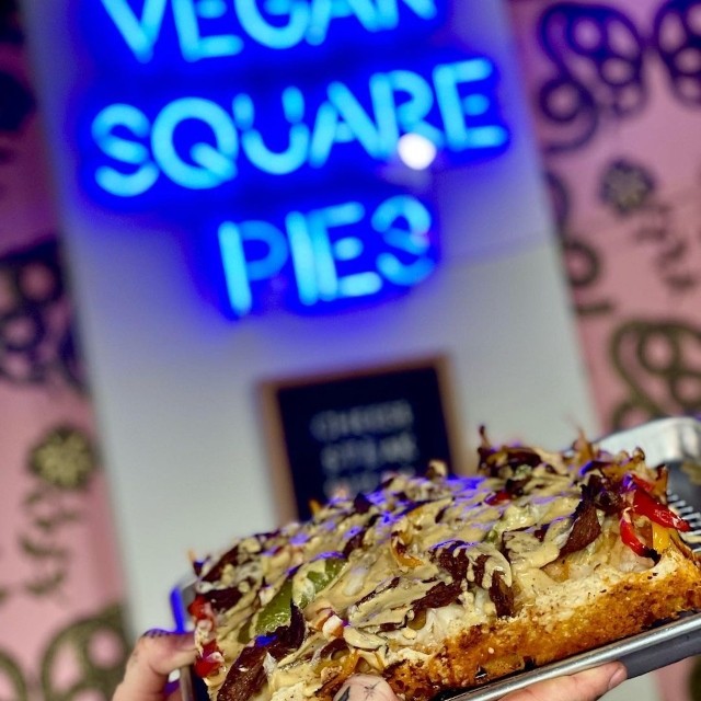 Hand holding a vegan square pizza slice in front of a blue custom neon 'Vegan Square Pies' sign on a pink patterned wall