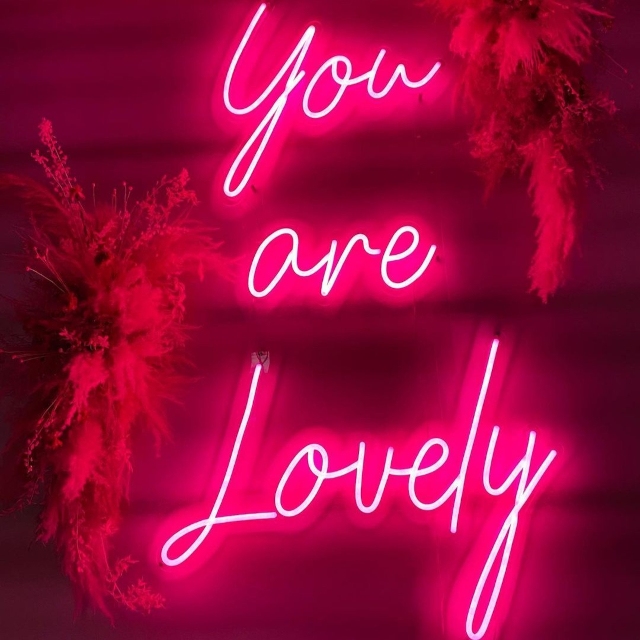 Pink custom neon 'You are Lovely' script sign on a wall flanked by pampas grass arrangements
