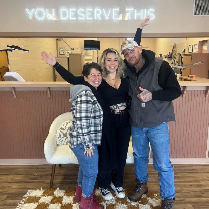 Three people posing for a selfie in front of a white Custom Neon 'You Deserve This' sign mounted on a wall